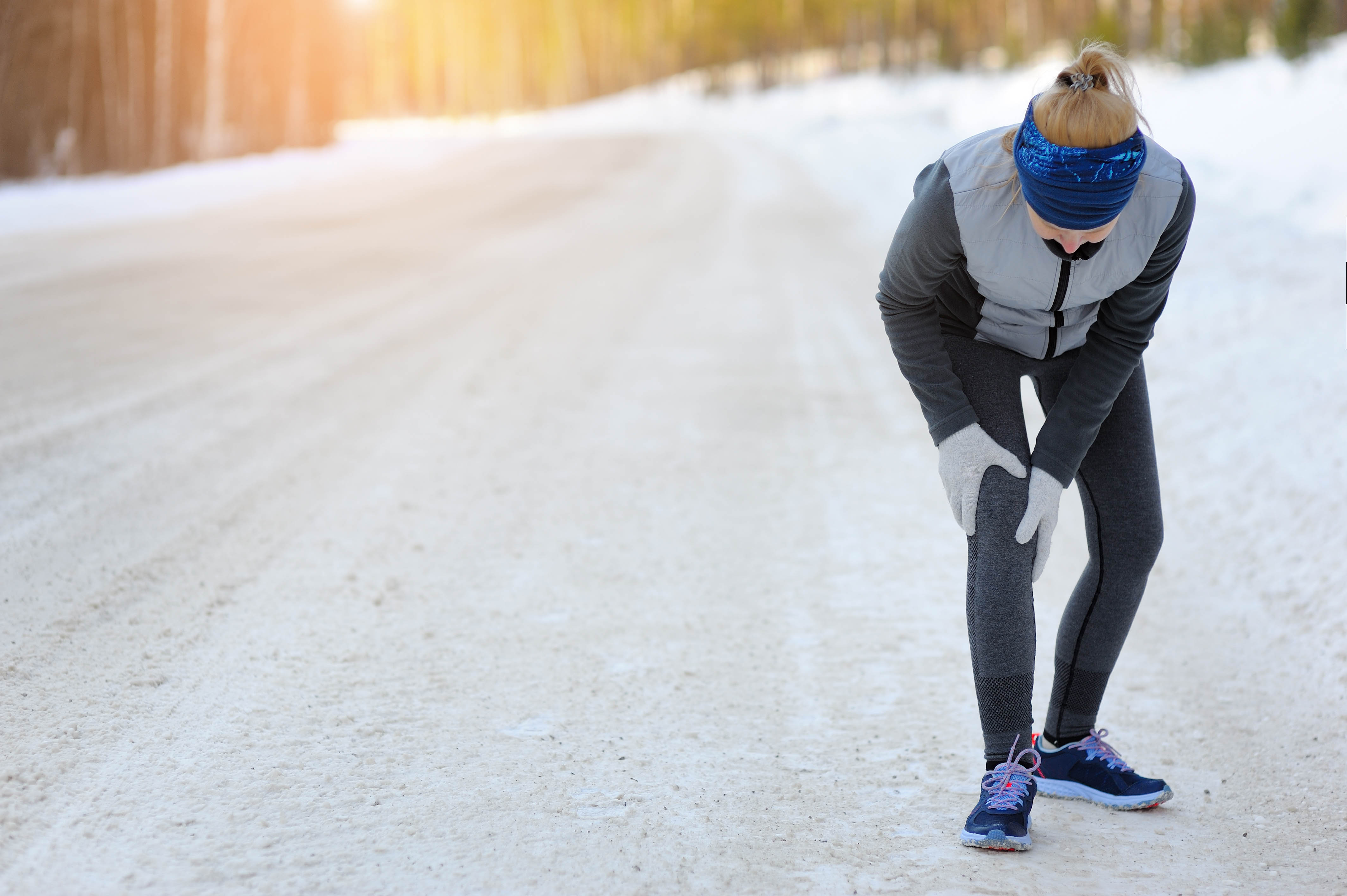 Woman wearing her winter clothing, bent over at the waist and holding her knee. Snow on the ground behind her